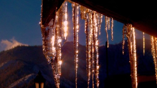 Icicles hang from a roof, reflecting sunlight against a backdrop of mountains and a clear blue sky.