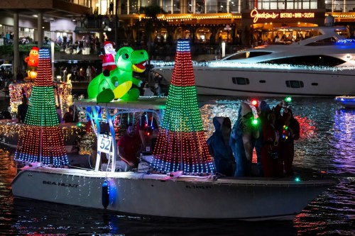 A festive boat decorated with colorful lights, featuring a dinosaur and Christmas trees, sailing on a waterway at night.