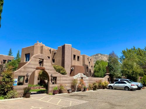 A southwestern-style building with adobe architecture, surrounded by greenery and parked cars under a clear blue sky.