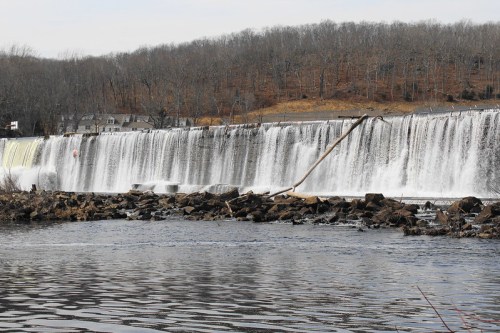 A waterfall cascades over a dam, surrounded by rocky shores and bare trees in the background.