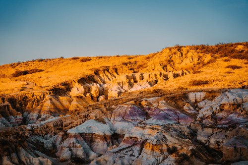 A colorful landscape of layered rock formations under a clear blue sky, illuminated by warm sunset light.