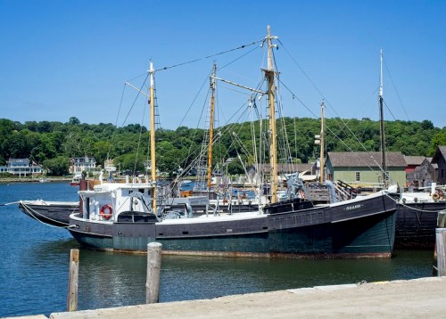 A historic fishing boat docked in a calm harbor, surrounded by green trees and quaint buildings under a clear blue sky.
