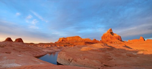 A panoramic view of red rock formations and a calm water body under a blue sky with scattered clouds.