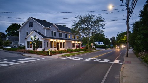 A quiet street scene at dusk featuring a charming building with a porch, streetlights, and a tree-lined sidewalk.