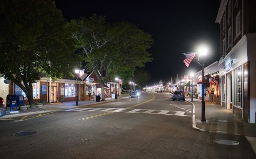A quiet street at night, lined with shops and trees, featuring streetlights and an American flag.