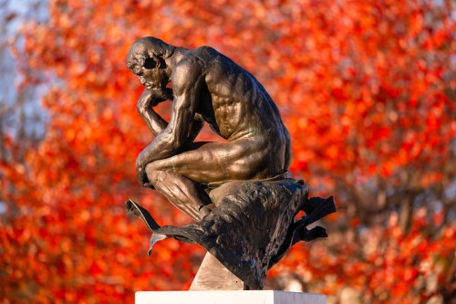 A bronze sculpture of a seated thinker against a backdrop of vibrant autumn leaves.