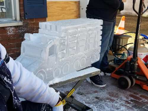 A partially carved ice sculpture of a truck, with a person working on it in the background.