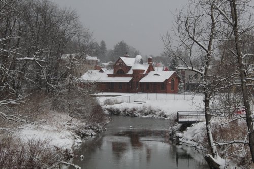 A snowy landscape featuring a red brick building by a river, surrounded by trees and a quiet, wintry scene.