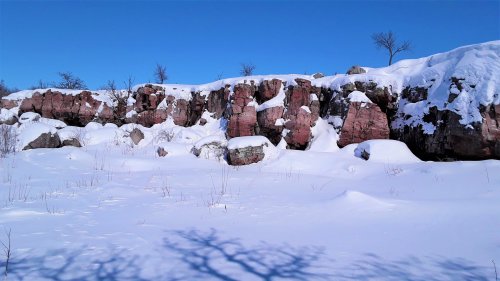 Snow-covered rocky cliff under a clear blue sky, with sparse trees and shadows on the snowy ground.
