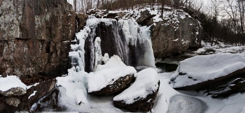 A frozen waterfall surrounded by snow-covered rocks and trees in a winter landscape.
