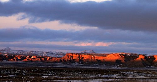 A panoramic view of rugged cliffs illuminated by sunset, with dramatic clouds overhead and a snowy landscape below.