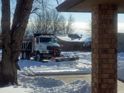 A snowplow truck clears a snowy residential street on a sunny winter day.