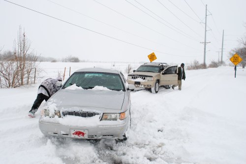 Two cars stuck in deep snow, with one person attempting to dig out a vehicle while another stands nearby.