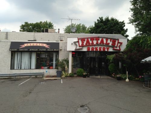 Exterior of Fattal's Bakery, featuring a sign and large windows, with a parking lot and trees in the background.