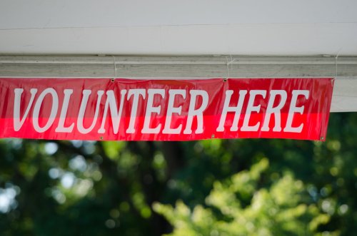 Red banner with white text that reads "VOLUNTEER HERE," hanging in a green outdoor setting.