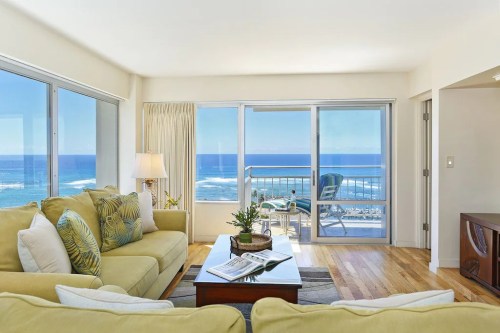 Bright living room with a yellow sofa, coffee table, and ocean view through large windows. Balcony visible in the background.
