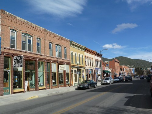 A charming street lined with historic brick buildings, shops, and parked cars, set against a backdrop of mountains and blue sky.