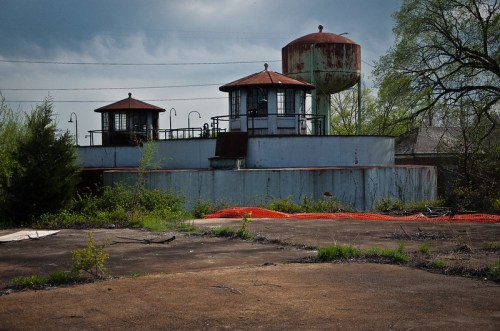 Abandoned industrial site with two water towers and overgrown vegetation under a cloudy sky.