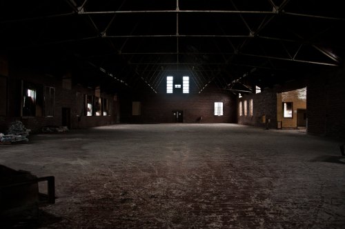 An empty, dimly lit industrial space with brick walls and large windows, featuring a dusty floor and high ceiling.