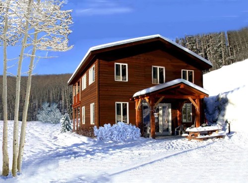 A cozy wooden house surrounded by snow, with a clear blue sky and trees in the background.