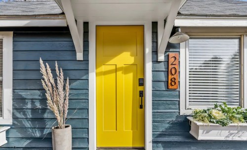 Bright yellow front door with modern design, flanked by a planter and house number 208 on a blue wooden exterior.