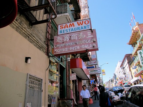 A bustling street scene in Chinatown featuring the Sam Wo Restaurant sign and pedestrians walking by.