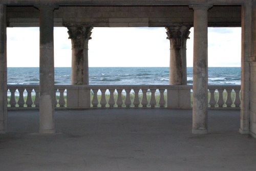 View of the ocean through columns and a balcony, with a cloudy sky and grassy area in the foreground.