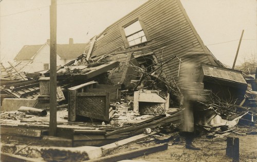 A collapsed house surrounded by debris, with a blurred figure walking past, captured in a historical photograph.