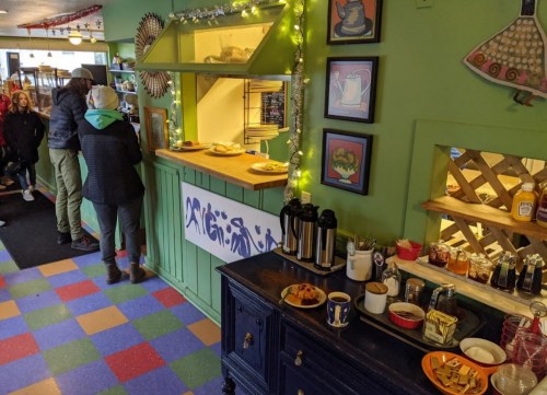 A cozy café interior with colorful flooring, a counter, and a table displaying pastries and drinks. Customers are chatting.