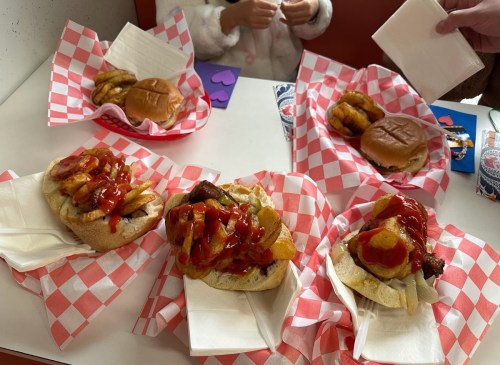 A table with four sandwiches in red-checkered baskets, topped with ketchup and fries, alongside napkins and hands.