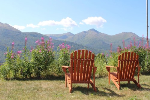 Two orange Adirondack chairs facing a scenic view of mountains and pink wildflowers under a clear blue sky.