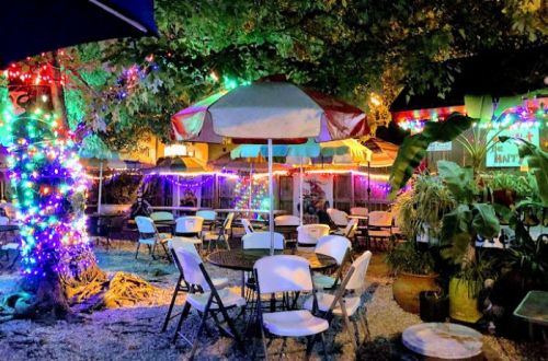 A vibrant outdoor dining area with colorful lights, umbrellas, and empty tables surrounded by greenery.