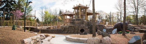 A playground featuring a wooden structure, rocks, and trees, set in a landscaped area with pathways and natural elements.