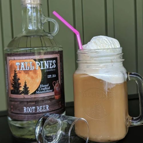 A mason jar filled with root beer topped with foam, next to a bottle of Tall Pines Distillery root beer moonshine.