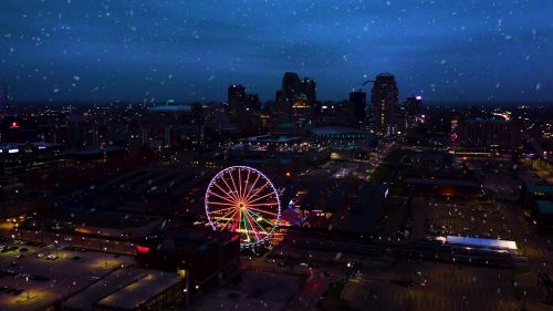 Aerial view of a city skyline at night with a brightly lit Ferris wheel and falling snow.