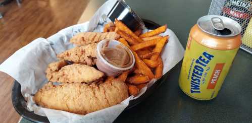 A plate of fried chicken tenders with sweet potato fries, dipping sauce, and a can of peach-flavored Twisted Tea.