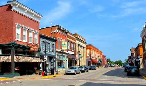 A charming small-town street lined with historic buildings, shops, and parked cars under a clear blue sky.