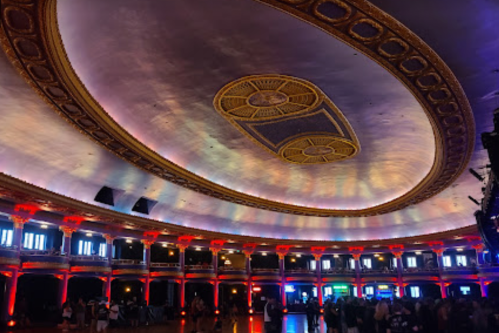 Interior of a grand hall with ornate ceiling, colorful lighting, and a crowd gathered on the floor.