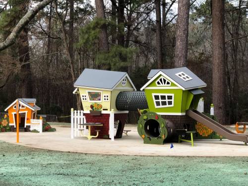 Colorful playground structure resembling houses, with slides and tunnels, surrounded by trees in a park setting.