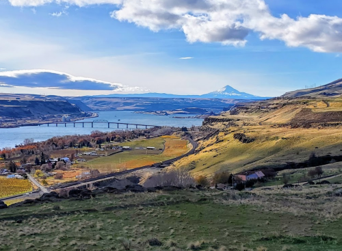 A scenic view of a river valley with a bridge, rolling hills, and a snow-capped mountain in the background.