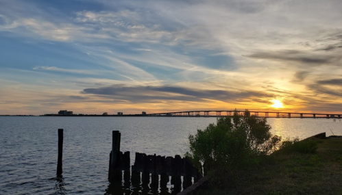 Sunset over a calm body of water, with a bridge in the distance and silhouettes of wooden posts in the foreground.