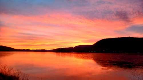 Vibrant sunset over a calm lake, with colorful skies reflecting on the water and hills in the background.