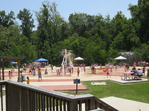 A vibrant splash pad filled with children playing in water, surrounded by trees and shaded areas on a sunny day.