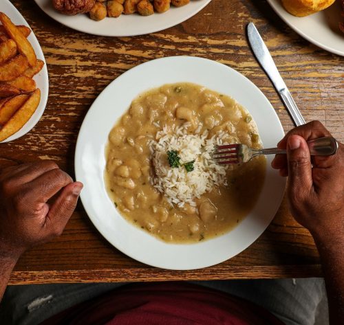 A plate of creamy white bean stew with rice, hands holding a fork, surrounded by various side dishes on a wooden table.