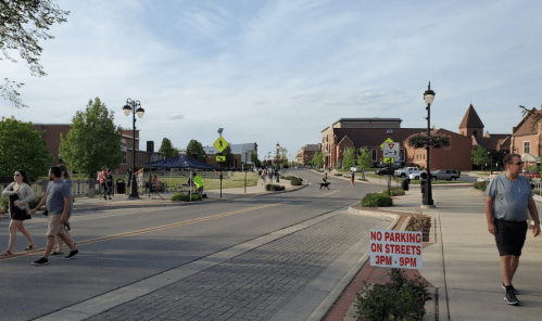 A busy street scene with pedestrians, shops, and a "No Parking" sign, under a clear sky.