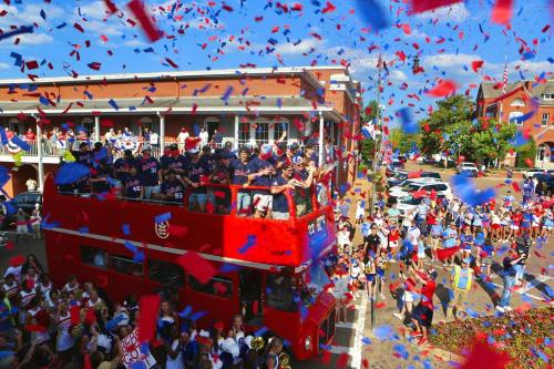 A double-decker bus in a parade, with people celebrating and confetti falling in red and blue.