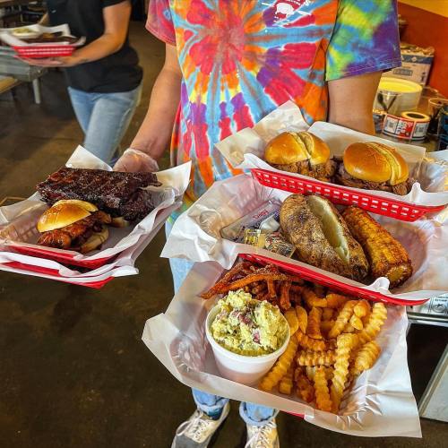 A person holds two trays of food featuring ribs, sandwiches, fries, and a baked potato in a casual dining setting.