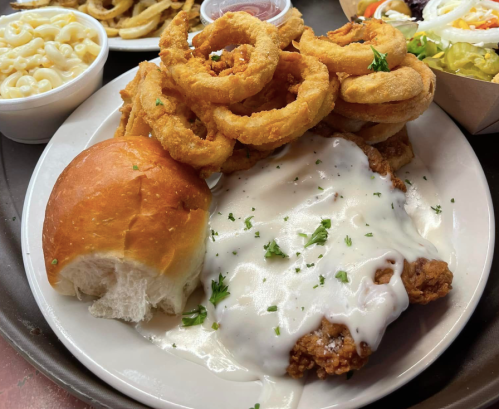 A plate of fried chicken topped with white gravy, served with onion rings, a roll, and a side of macaroni and cheese.