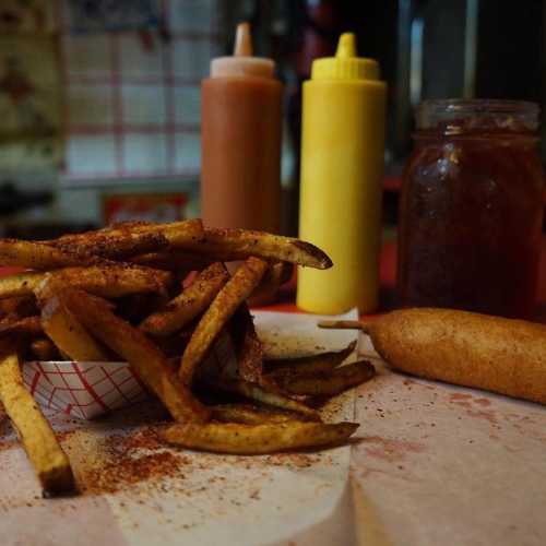 A serving of seasoned fries in a paper tray, with various condiments in squeeze bottles and a jar nearby.