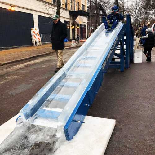 A large ice slide set up on a street, with a person standing nearby and a few people in the background.